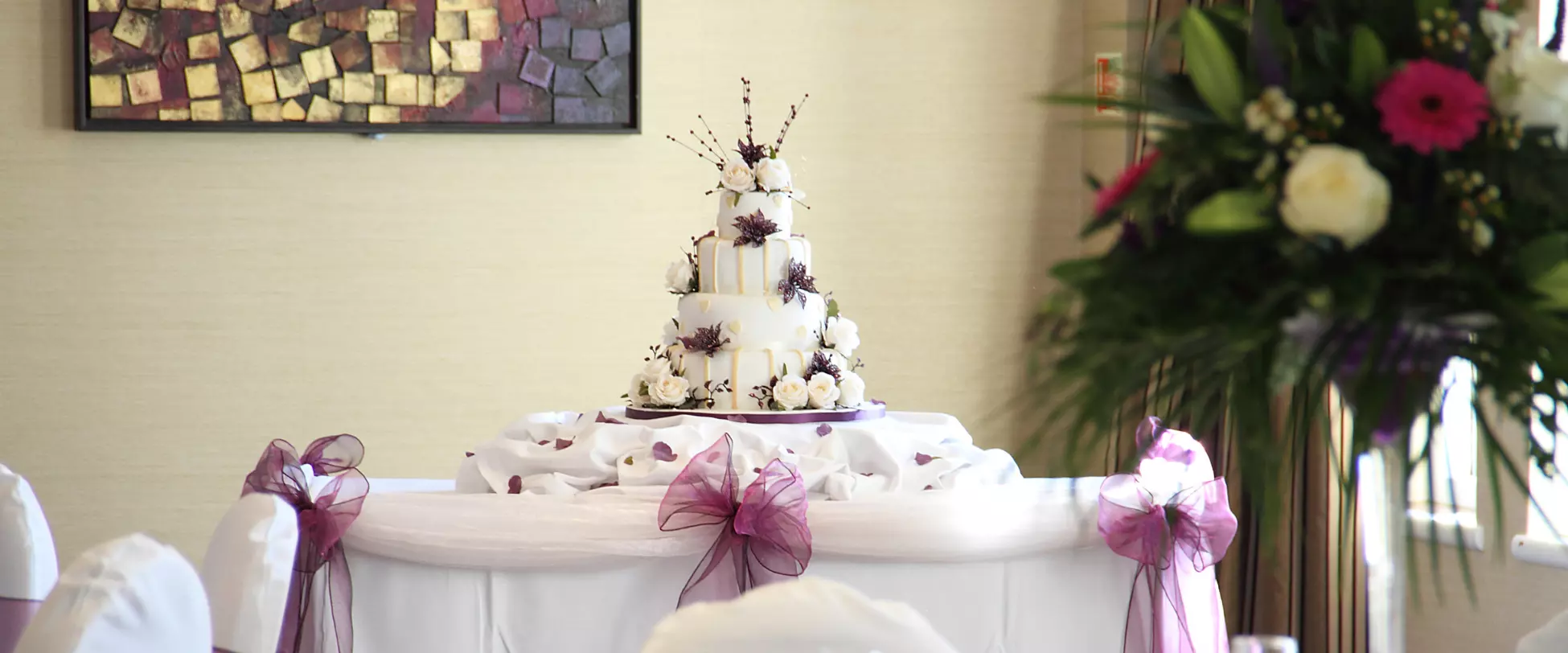 A three-tier wedding cake decorated with purple and white flowers sits on a table in the centre of the image. There are purple bows on the table and purple and white flowers to the right of the image.