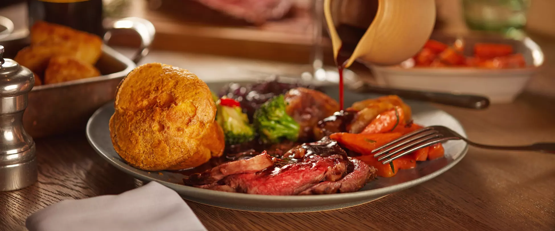 A plate of food on a wooden table. The plate has a roast beef, carrots, broccoli, roasted potatoes, a crispy Yorkshire pudding and someone is pouring gravy onto the food from a gravy jug..