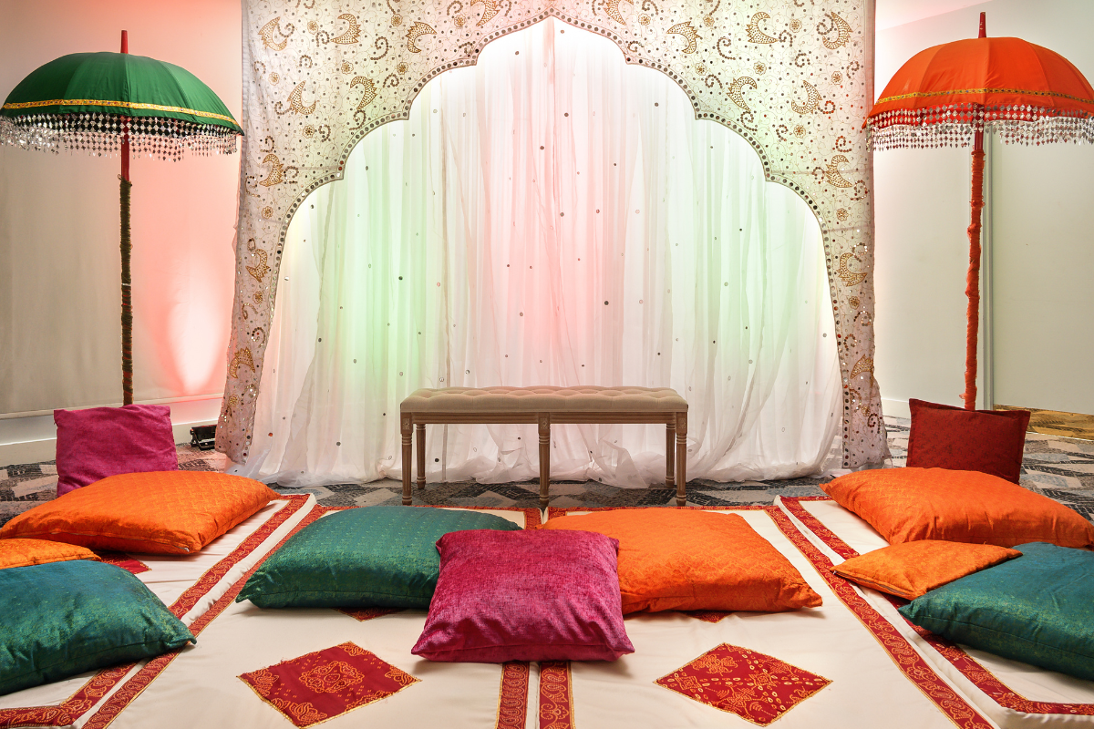 Green, orange and pink cushions surround a two seater bench. There are colourful umbrellas to either side, and behind the bench is a backdrop of white cloth embroidered with an intricate gold arch..