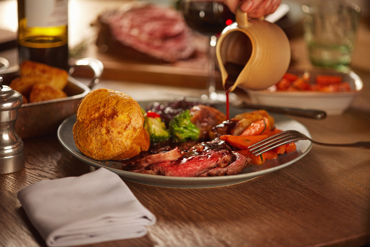 A plate of food on a wooden table. The plate has a roast beef, carrots, broccoli, roasted potatoes, a crispy Yorkshire pudding and someone is pouring gravy onto the food from a gravy jug..